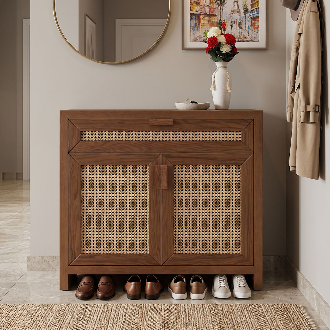 A wooden entryway cabinet with woven cane panels stands against a beige wall in a styled interior. A white vase with flowers sits on top, and several brown and white shoes are arranged underneath.